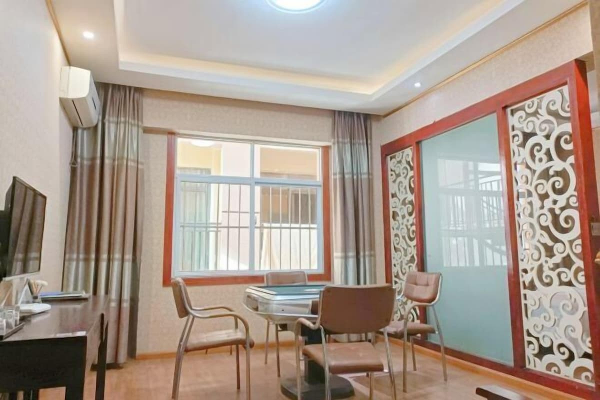 A hotel room with a dining table, four chairs, a television, and a large window with curtains.