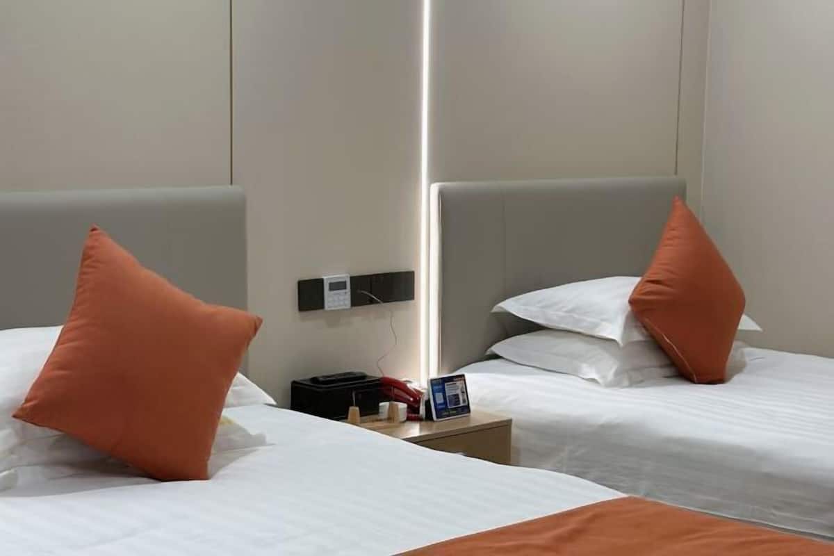 A hotel room with two beds, each with white bedding and an orange pillow. There is a bedside table with a phone, a small lamp, and a magazine.