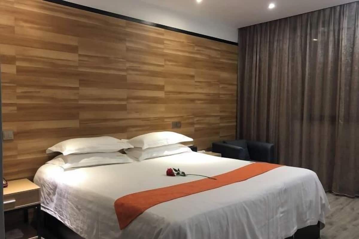 A hotel room with a wooden headboard, a bed with white bedding and an orange throw, a desk with a chair, and a large window with curtains.