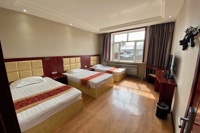 A hotel room with three beds, a desk, a chair, a radiator, and a window with curtains.