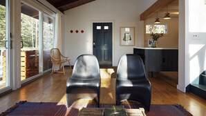 A spacious room with a large sliding glass door leading to a deck, a dark wooden door, and a kitchen area with a window and pendant lights.