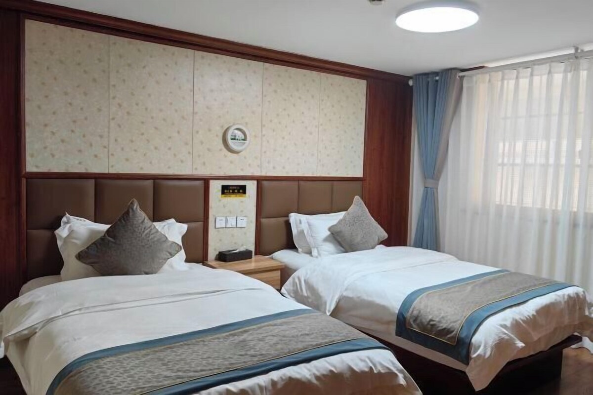 Una habitación de hotel con dos camas, un cabecero con un reloj, una mesita de noche y una ventana con cortinas.