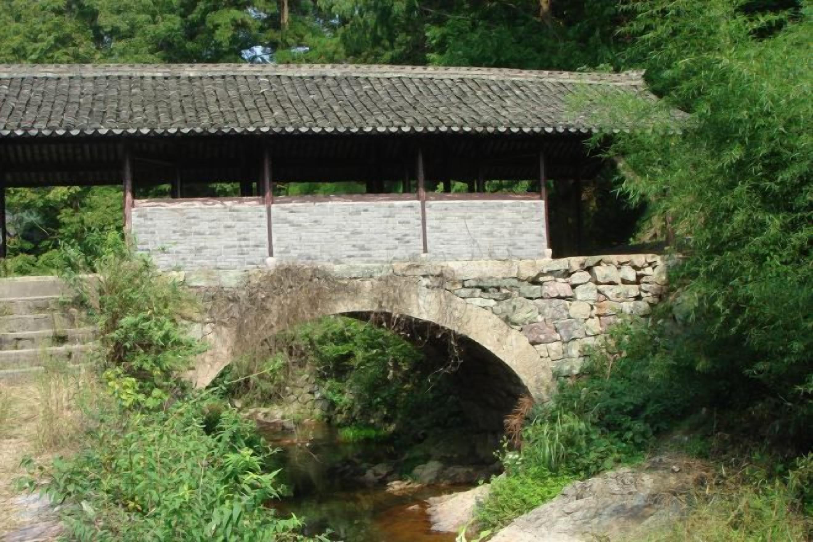 A stone bridge with a tiled roof over a stream, surrounded by greenery.