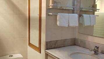 Un baño con inodoro, lavamanos y espejo. Toallas cuelgan de un perchero.