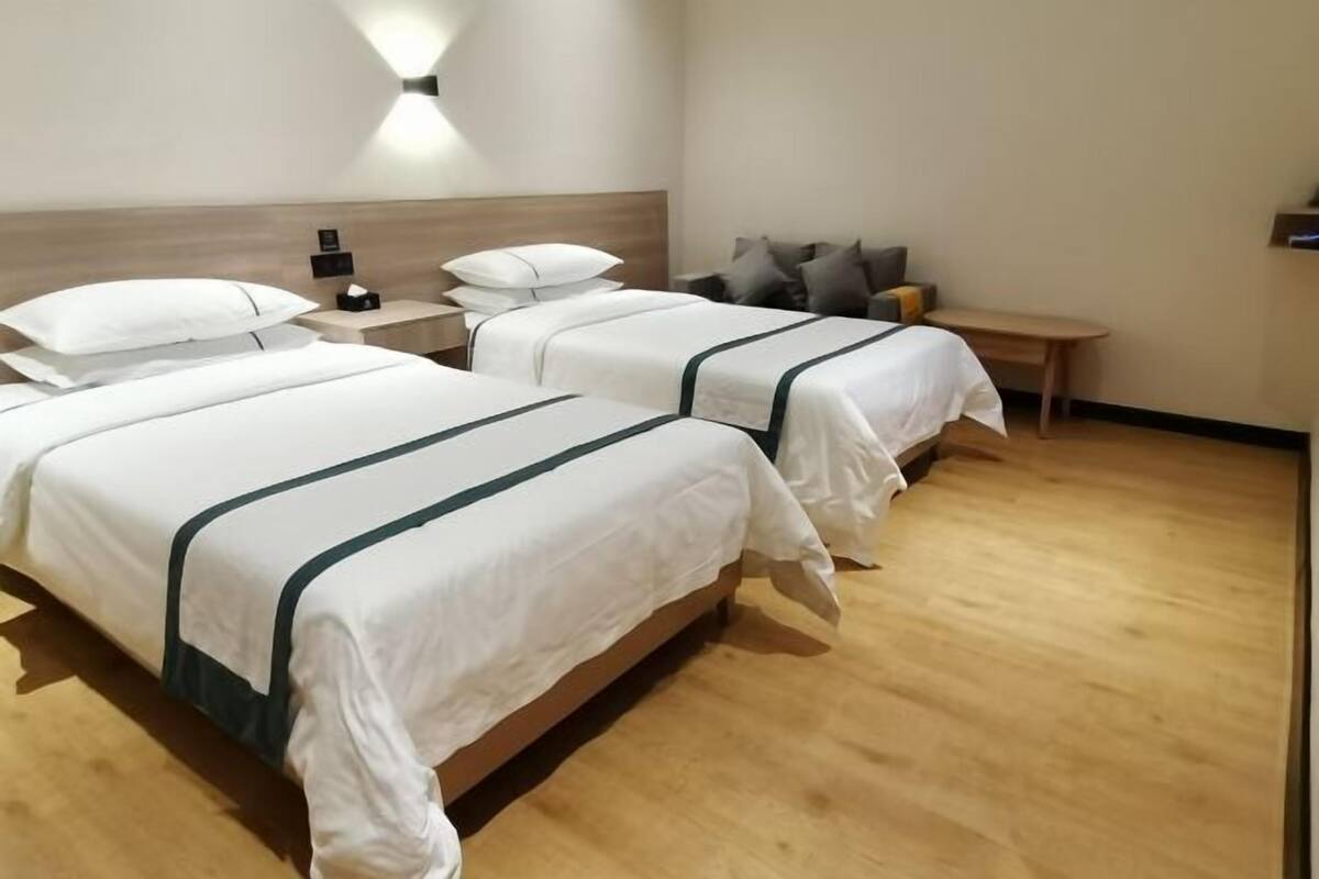 A hotel room with two beds, a bedside table with a lamp, a bench, and a wall-mounted light fixture.