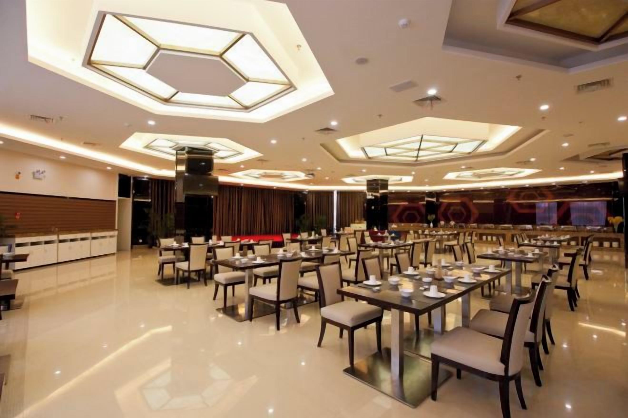 Un vaste espace dédié à la salle à manger, avec de nombreuses tables et chaises, un plafond haut orné de luminaires aux formes géométriques, et une scène en arrière-plan.