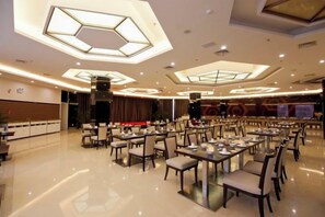 A spacious dining area with multiple tables and chairs, high ceiling with geometric light fixtures, and a stage in the background.