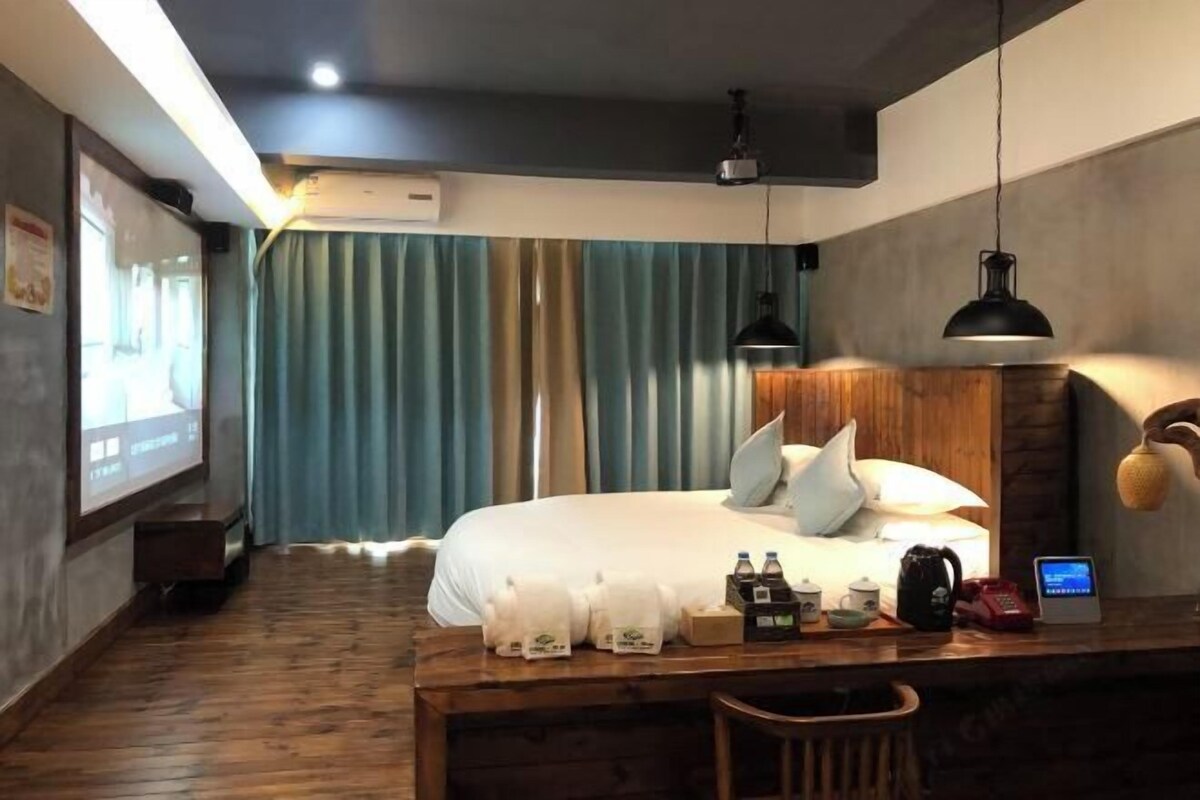 A hotel room with a bed, a wooden desk with amenities, a projector screen, and a large window with curtains.