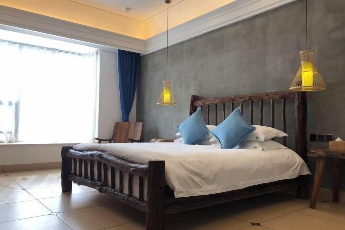 A bed with a wooden headboard, two blue pillows, and a white bedspread in a room with a large window, a chair, and two hanging lamps.