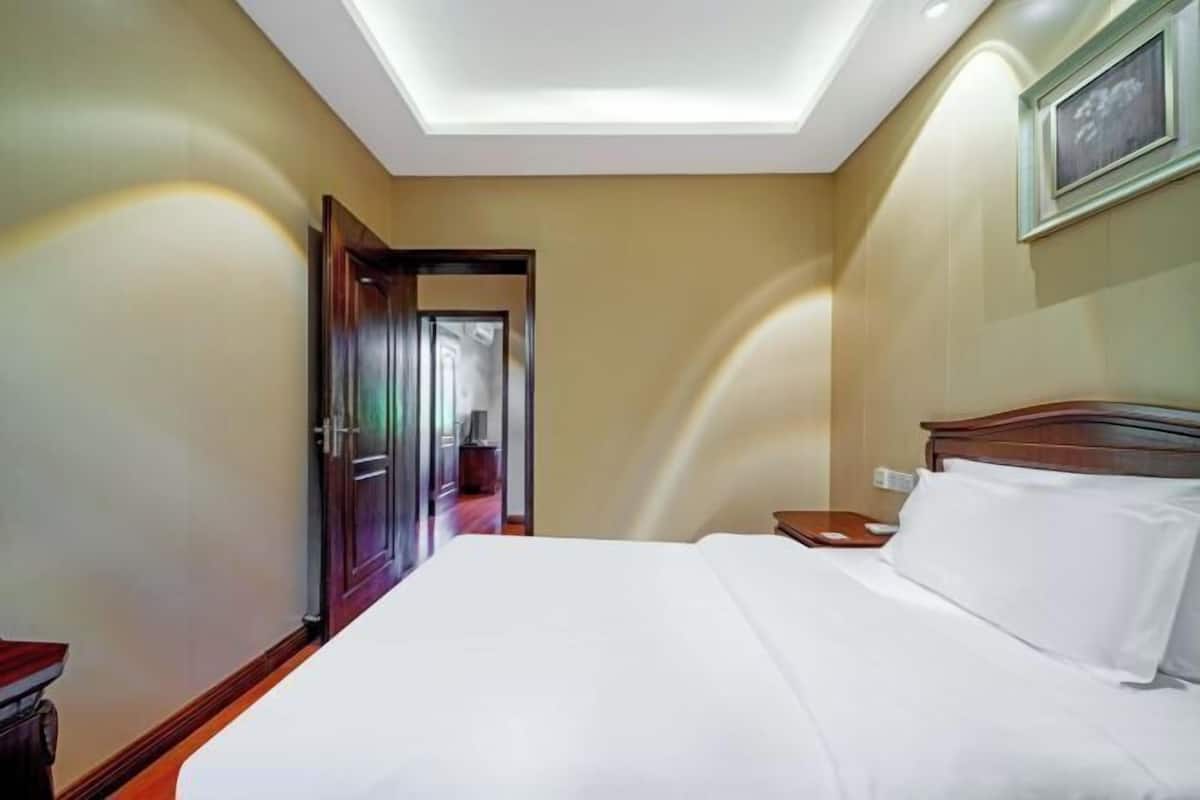 A hotel room with a large bed, a wooden nightstand, a ceiling light, and a door leading to another room.