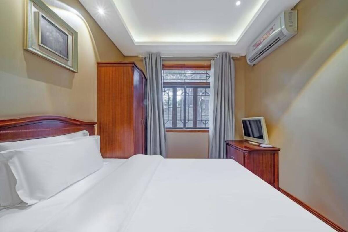 A hotel room with a bed, a nightstand, an air conditioner, a window with curtains, and a framed picture on the wall.