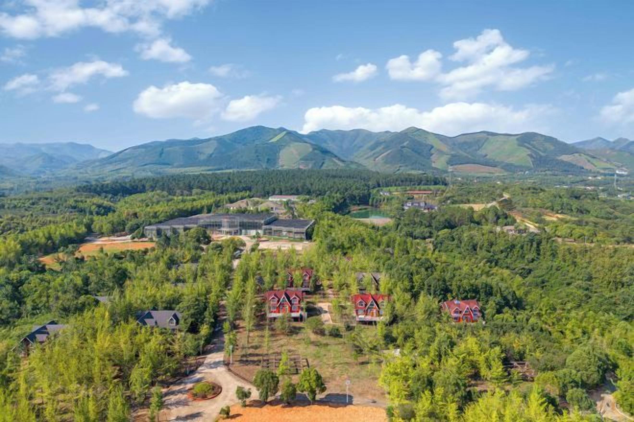 A rural landscape with a large building and several red-roofed houses amidst a forested area with mountains in the background.