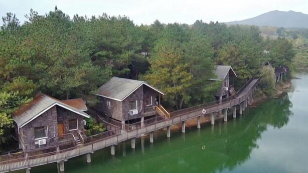 Una casa flotante de madera con cubierta y escaleras, conectada por un puente a una zona boscosa a orillas del agua.