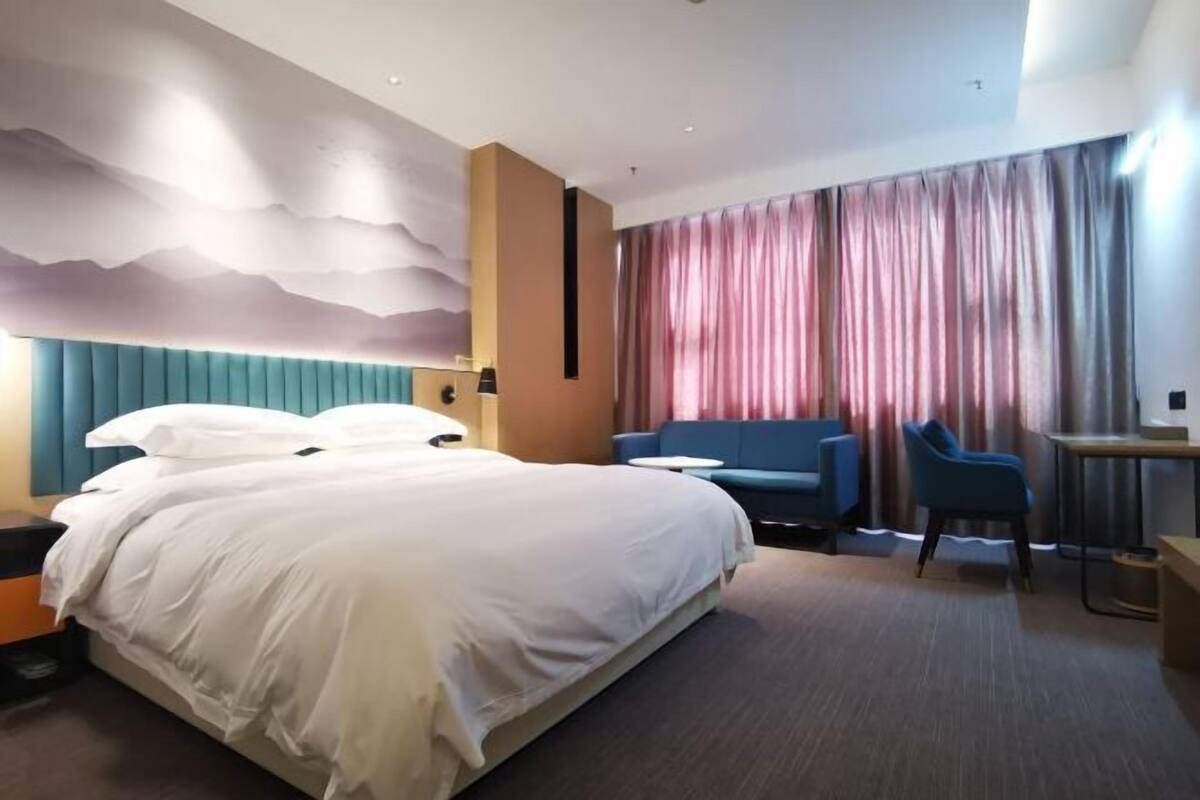 A hotel room with a large bed, a blue sofa, a small table, and a view of the city through curtains.