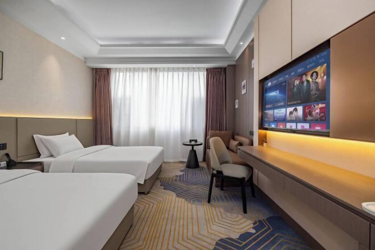 A hotel room with two beds, a desk, a TV, and a large window with curtains.