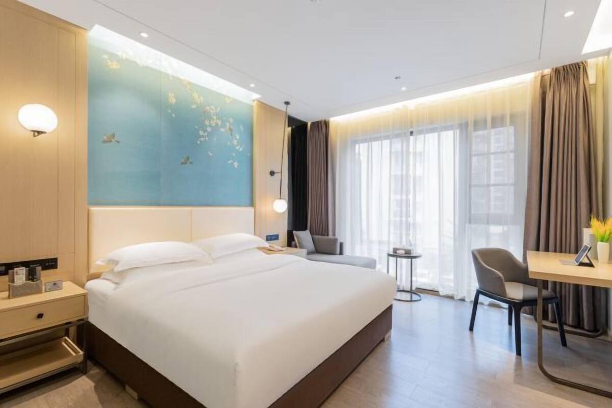 A modern hotel room with a large bed, a desk, and a chair. There is a small table and a lamp on the desk.