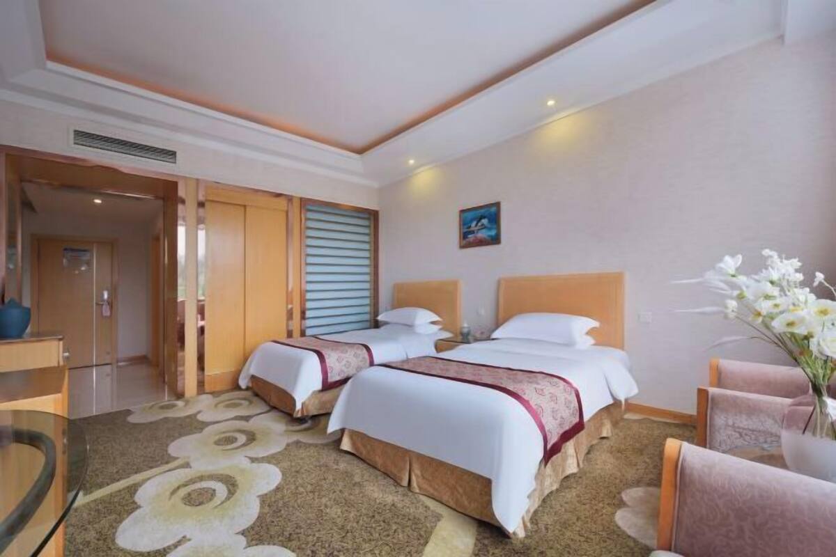 A hotel room with two beds, a sitting area with a chair, a small table, and a decorative vase with flowers.