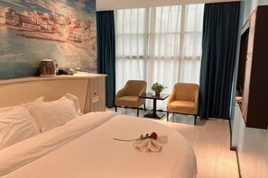 Habitación de hotel con una cama, un escritorio, dos sillas, una ventana con cortinas y un cuadro en la pared.