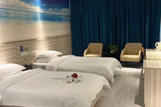 Ein Hotelzimmer mit zwei Betten, einem Schreibtisch, zwei Stühlen, einem Nachttisch und einem Wandbild mit einer Strandansicht.