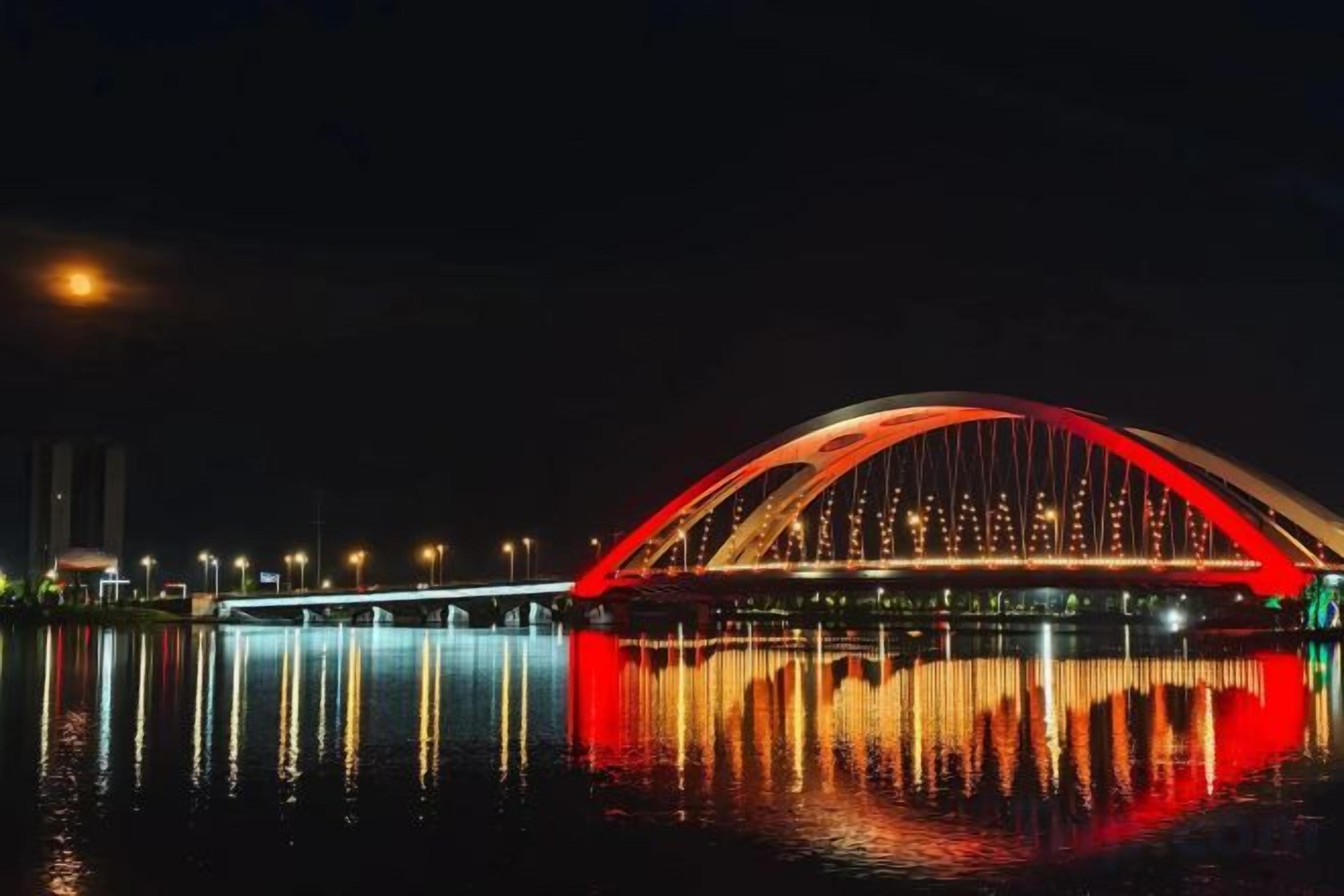 Vista nocturna de un puente con un arco rojo y luces que se reflejan en el agua.