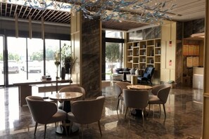 A hotel lobby with a marble floor, round tables, and chairs, a large window with a view of the street, and a bookshelf with various items. - Boao Hotel (Yancheng)