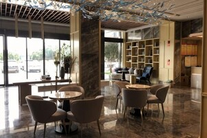 Un vestíbulo de hotel con piso de mármol, mesas y sillas redondas, un gran ventanal con vista a la calle, y una estantería con diversos objetos.
