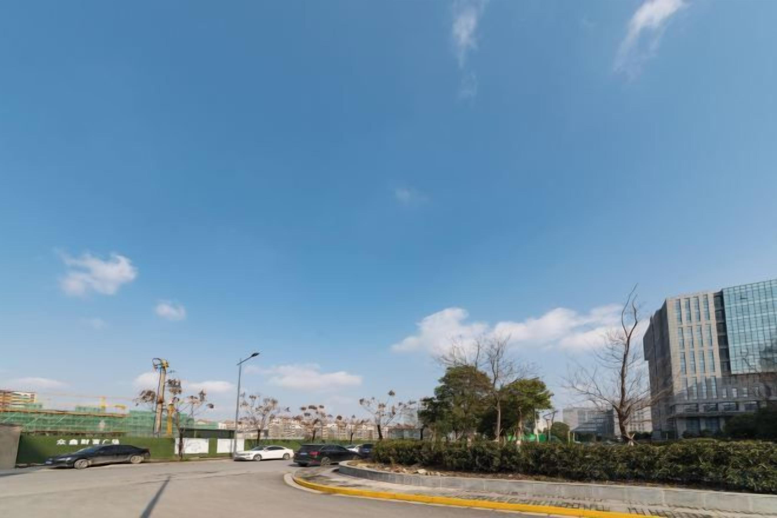 Klarer Himmel mit vereinzelten Wolken, eine Straße mit gelber Bordsteinkante und Gebäude im Hintergrund.