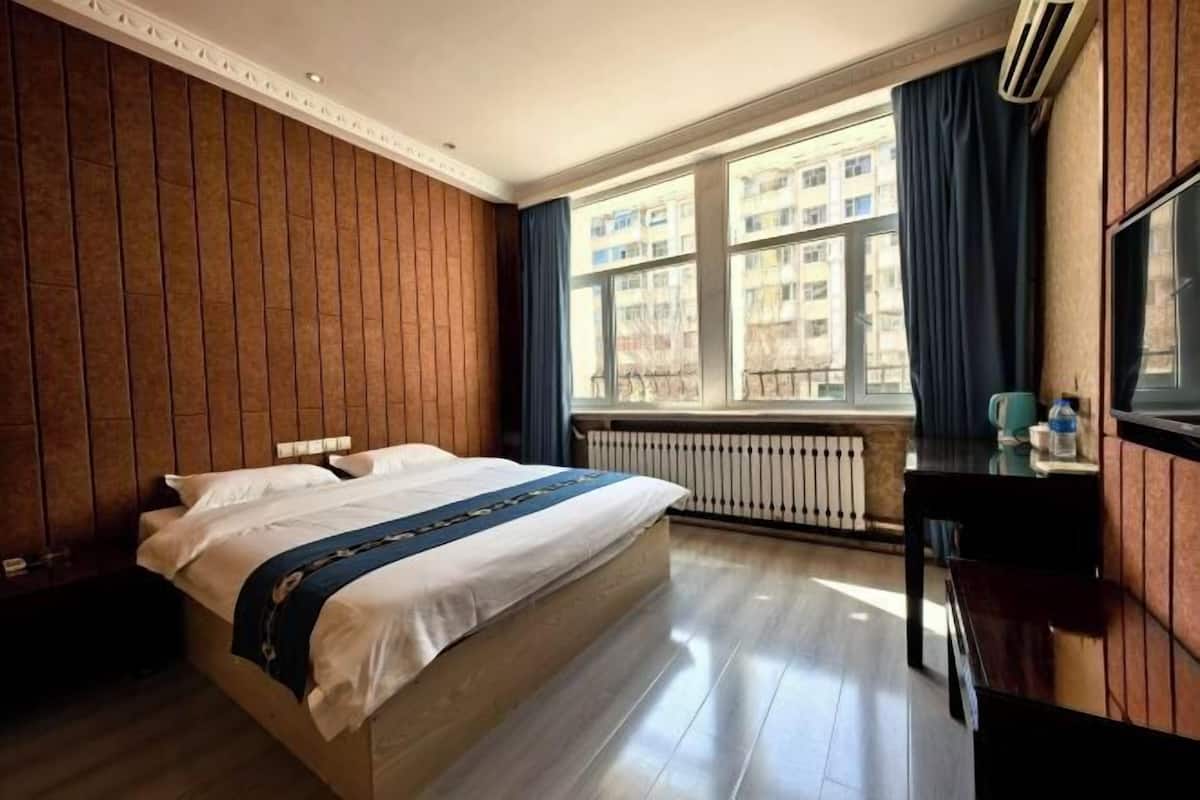 Une chambre d’hôtel avec un grand lit, une fenêtre donnant sur des immeubles, un radiateur et un bureau avec une chaise.