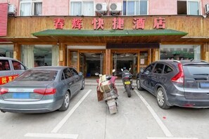 Entrada de hotel con toldo verde y un letrero arriba. Dos coches estacionados al frente y una motocicleta con un paquete de entrega.