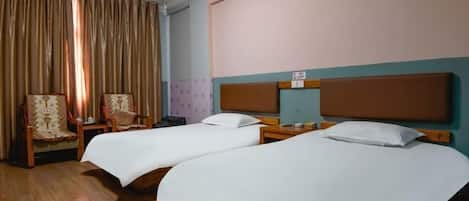 A hotel room with two single beds, a wooden floor, a large window with curtains, and a small table with a chair.