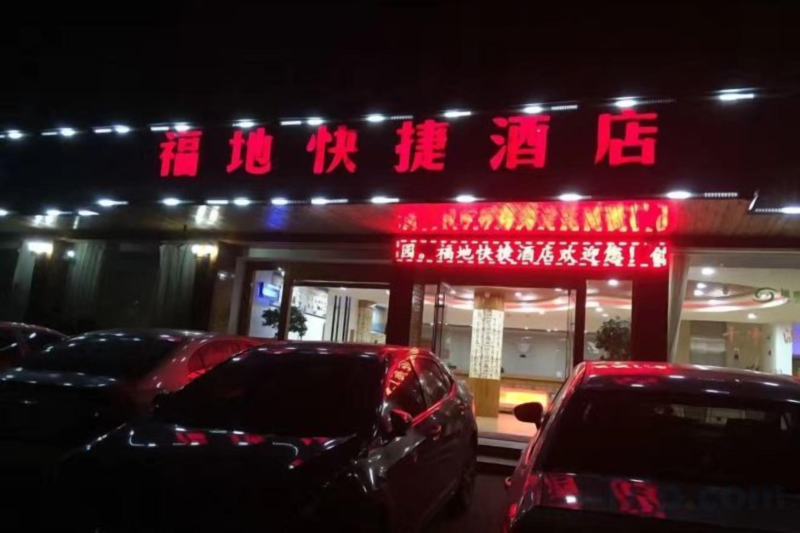 L’entrée d’un hôtel, avec une enseigne rouge, un parking et un bâtiment dont les fenêtres sont illuminées.