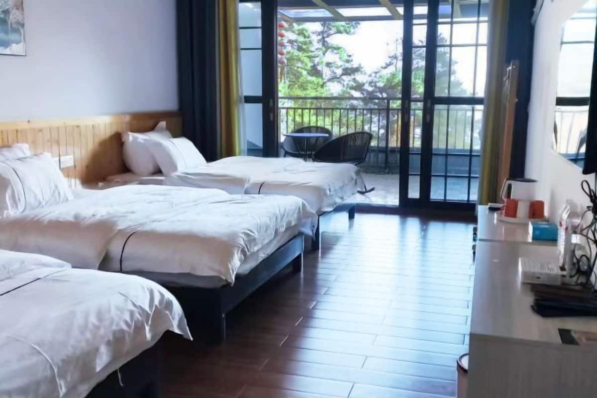 A hotel room with three beds, a wooden floor, a balcony with a chair, and a desk with a phone and a cup.