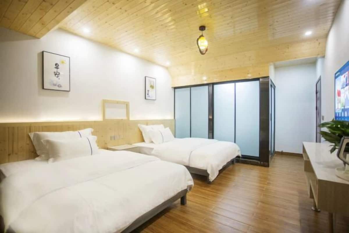 A hotel room with two beds, wooden flooring, a desk, and a large window.