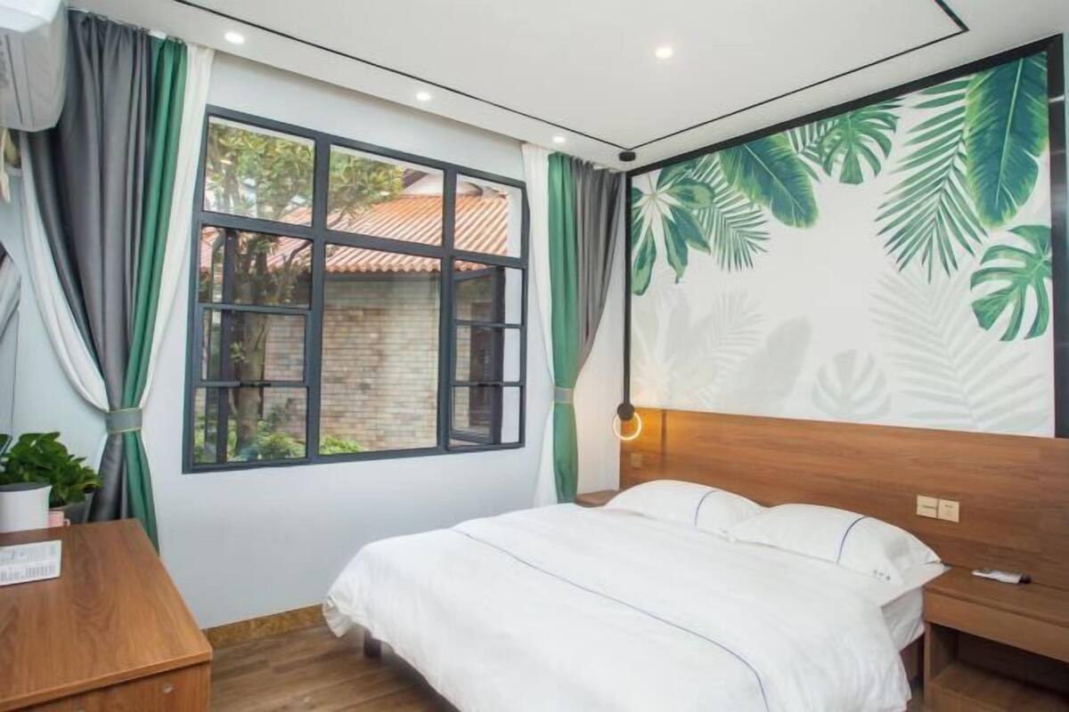 A bedroom with a bed, a wooden headboard, a window with curtains, a wall mural, and a bedside table with a plant.