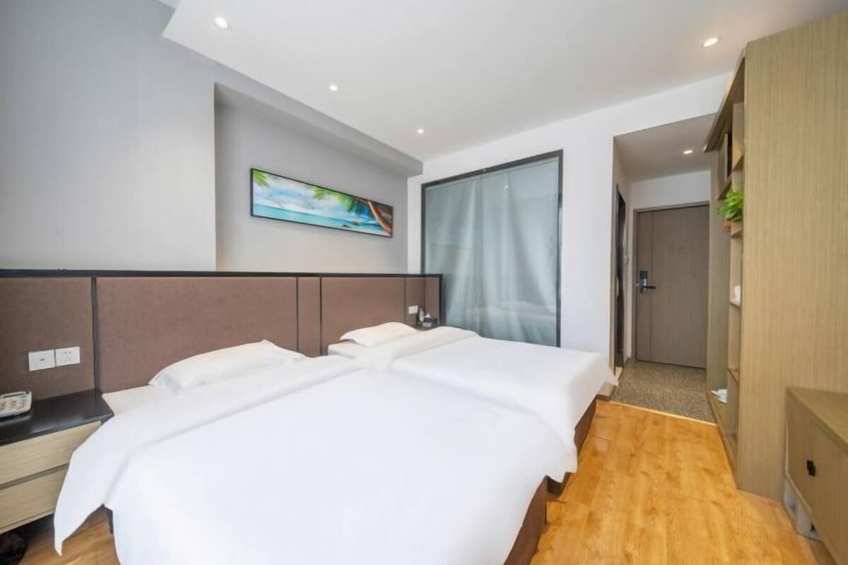 A hotel room with two beds, a wooden floor, a painting on the wall, and a large window with curtains.