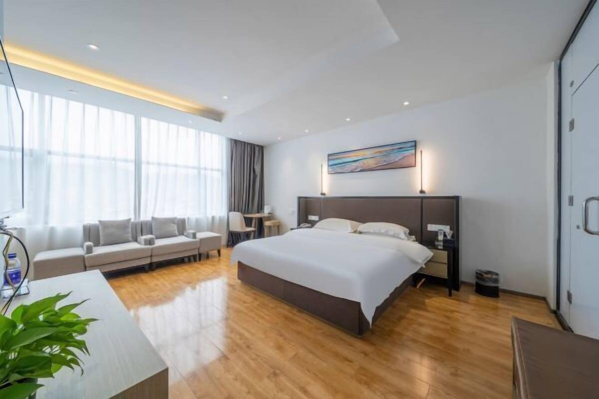 A modern hotel room with a large bed, a sofa, a desk, and a large window with curtains.