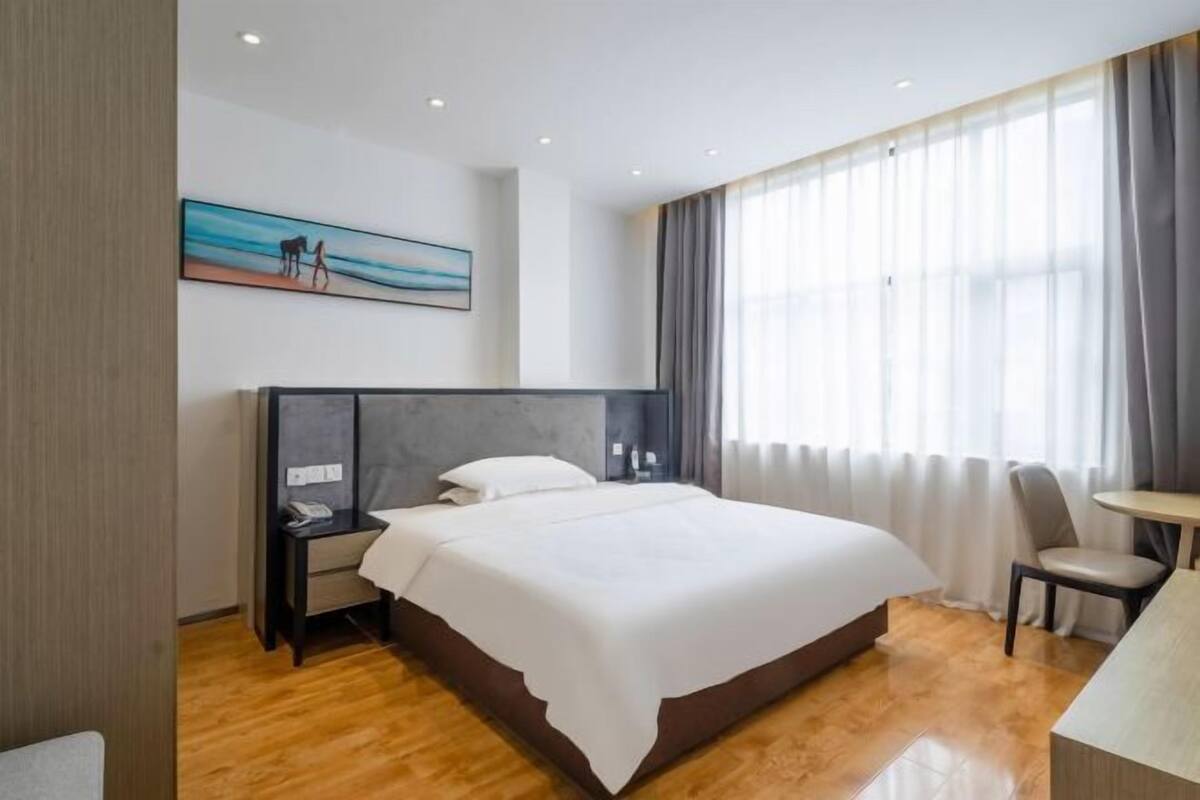 A hotel room with a large bed, a bedside table, a chair, a desk, and a window with curtains.