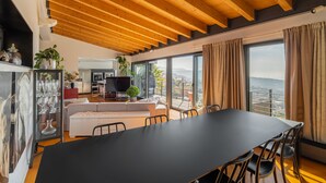A modern living room with a large dining table, a sofa, and a TV. There are large windows with curtains, a balcony, and a view of the city. - Amazing 4 bedroom Villa with sea view pool (Alella Parc)