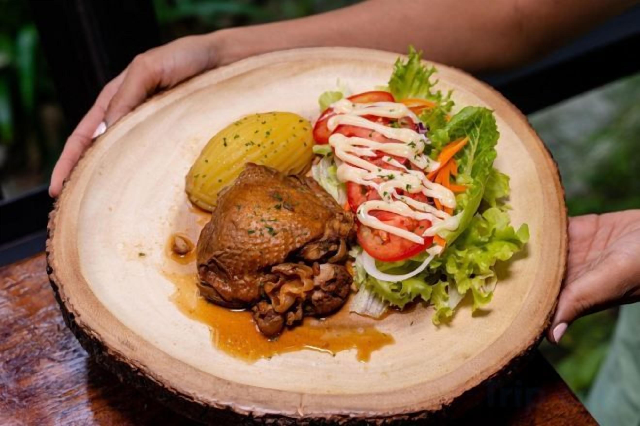 Um prato de madeira com frango assado, batata e salada com molho cremoso.