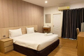 A hotel room with a wooden headboard, a single bed, a bedside table, a small bedside cabinet, a mirror, a sink, a door, and a curtain. - Crystal Resort (Nakhon Ratchasima)
