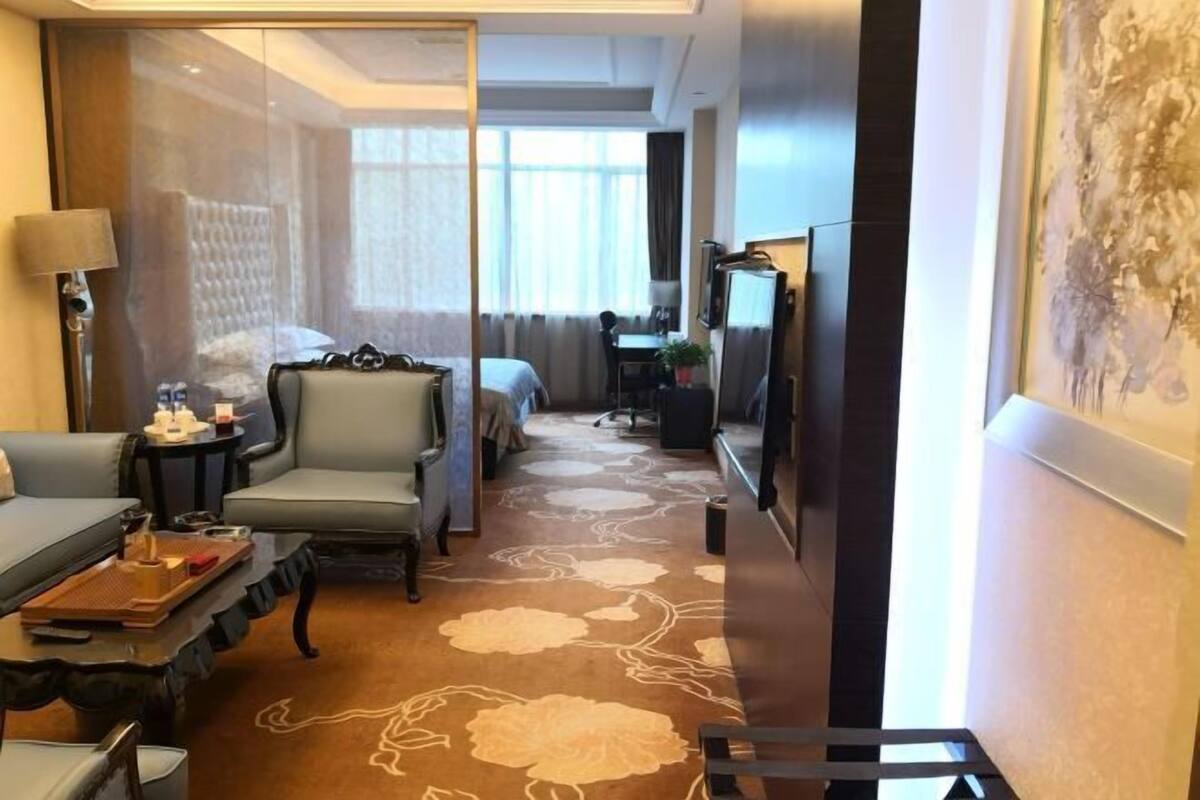 A hotel room with a sofa, a chair, a small table with a tray, a bed, a desk with a chair, a television, and a large painting on the wall.