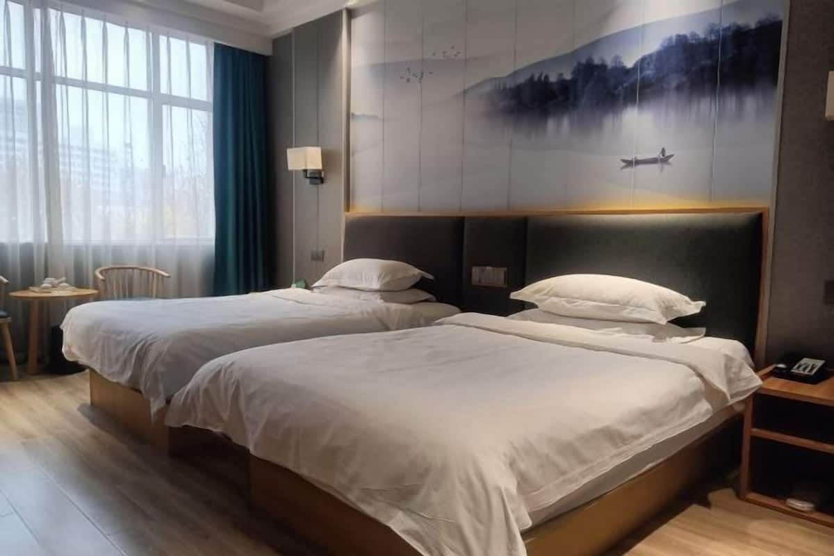 A hotel room with two beds, a large window with curtains, a painting of a lake scene, and a small table with a chair.