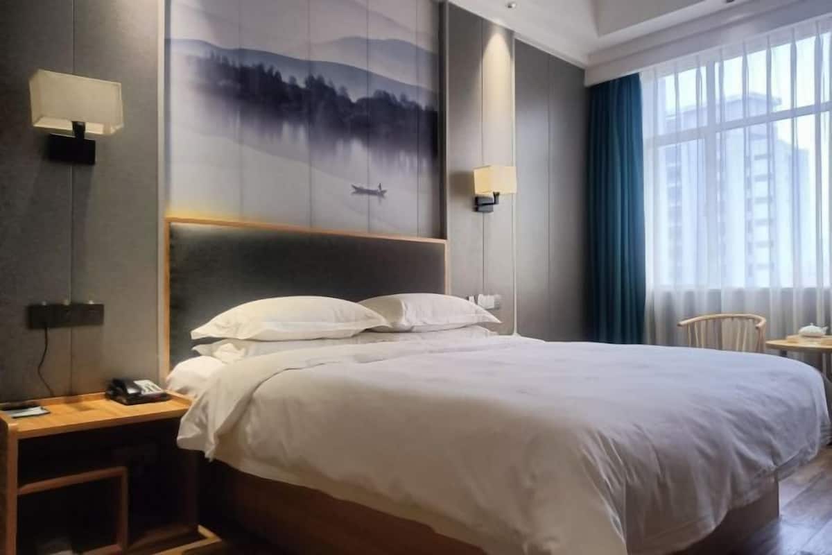 A hotel room with a large bed, a nightstand with a phone, a wall-mounted lamp, and a window with curtains.