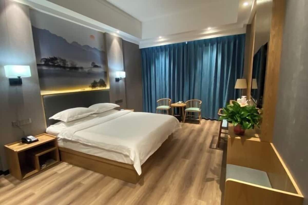 A hotel room with a bed, bedside tables, a desk with chairs, a desk lamp, a potted plant, and a mural on the wall.