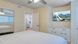 A bedroom with a bed, a ceiling fan, a door leading to a bathroom, and a wall-mounted TV displaying a sailboat on the water. - Monthly Only - Remodeled Lower Unit Just One House To The Sand With AC (Newport Beach)