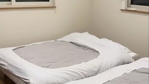 Una cama individual con ropa de cama blanca y gris en una habitación con paredes lisas y una ventana.
