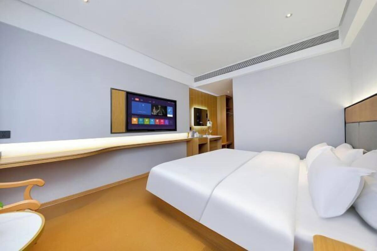 A modern hotel room with a large bed, a bedside table, a flat-screen TV mounted on the wall, and a wooden headboard.