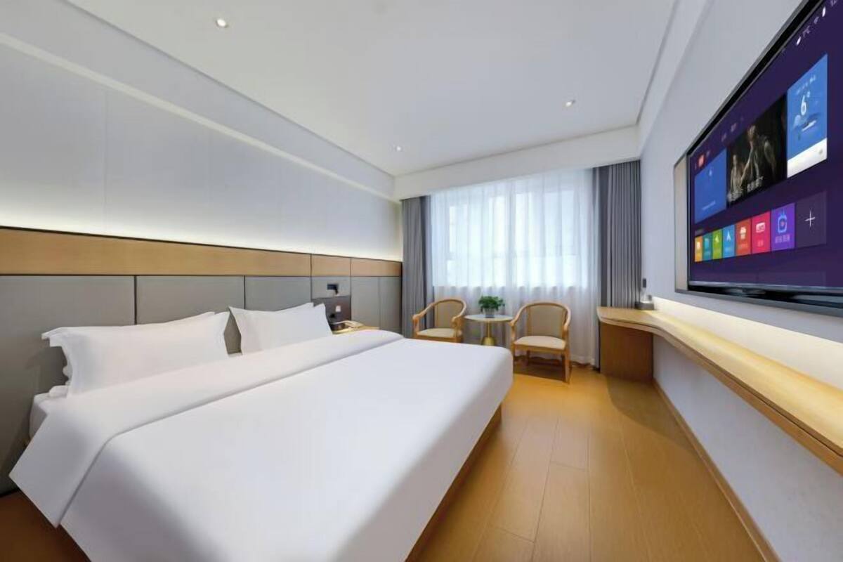 A hotel room with a large bed, a TV mounted on the wall, a small table with chairs, and a window with curtains.