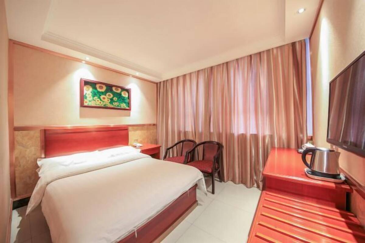 A hotel room with a bed, a desk with a kettle, a chair, a TV, and a painting on the wall.