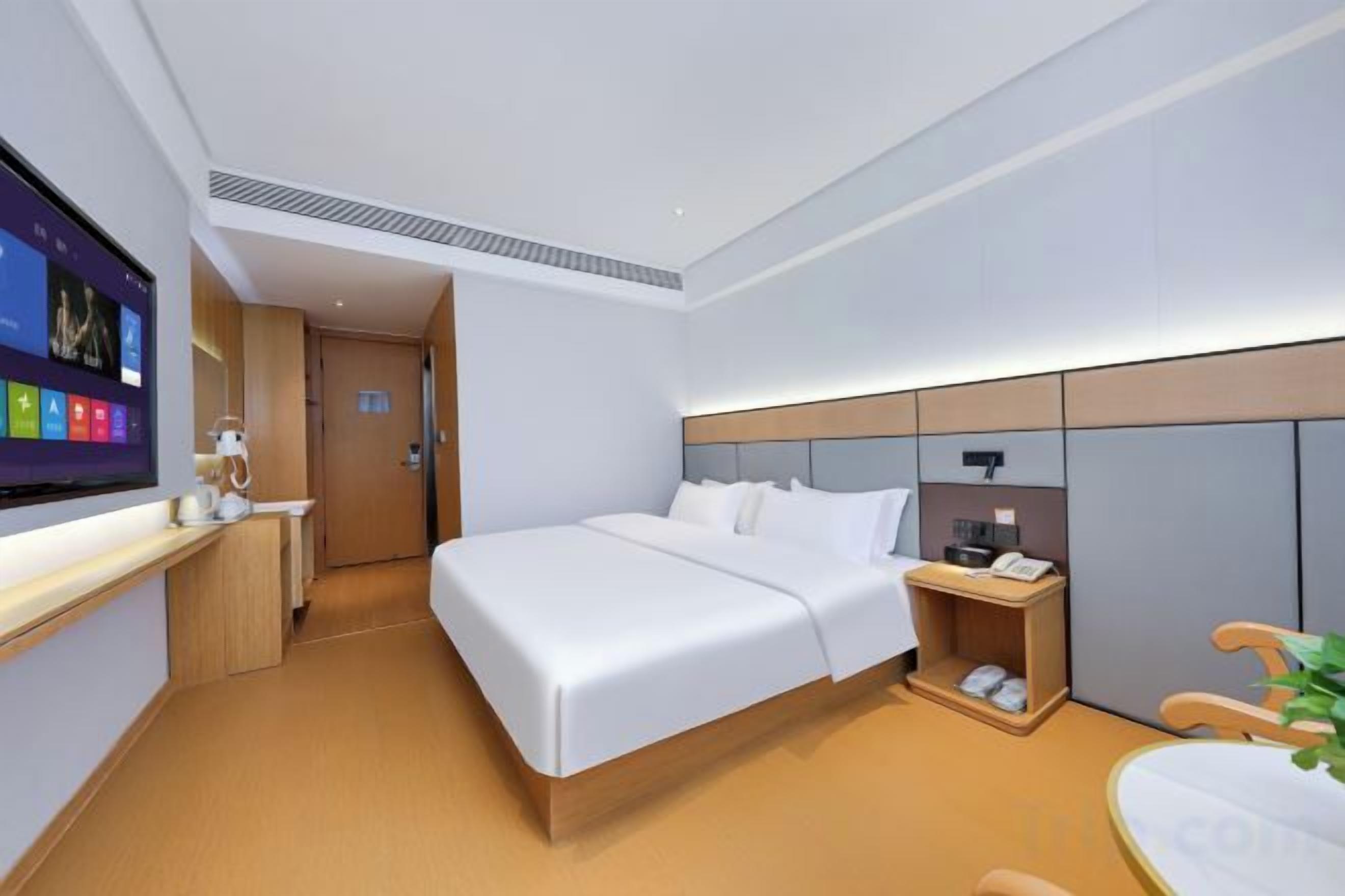 A modern hotel room with a bed, bedside table, TV, and a desk with a chair.